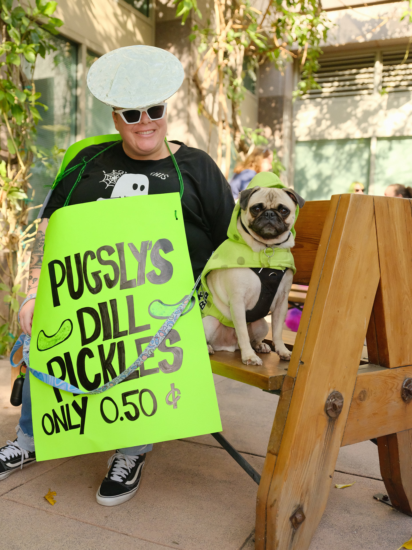 A pug dressed as a dill pickle and his owner sit on a bench.